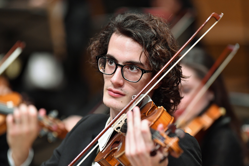 A man with dark curly hair, wearing glasses, playing the violin, wearing a smart suit.